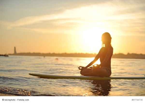 Eine junge Frau sitzt meditierend auf einem Surfbrett an einem Strand mit untergehender Sonne