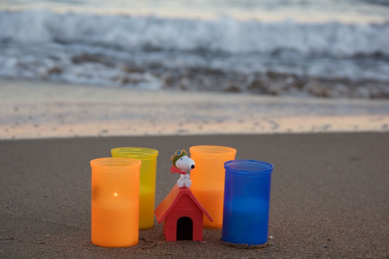 4 Kerzen um einen Snoopy auf seiner roten Hütte stehen an einem Strand im Hintergrund die Wellen