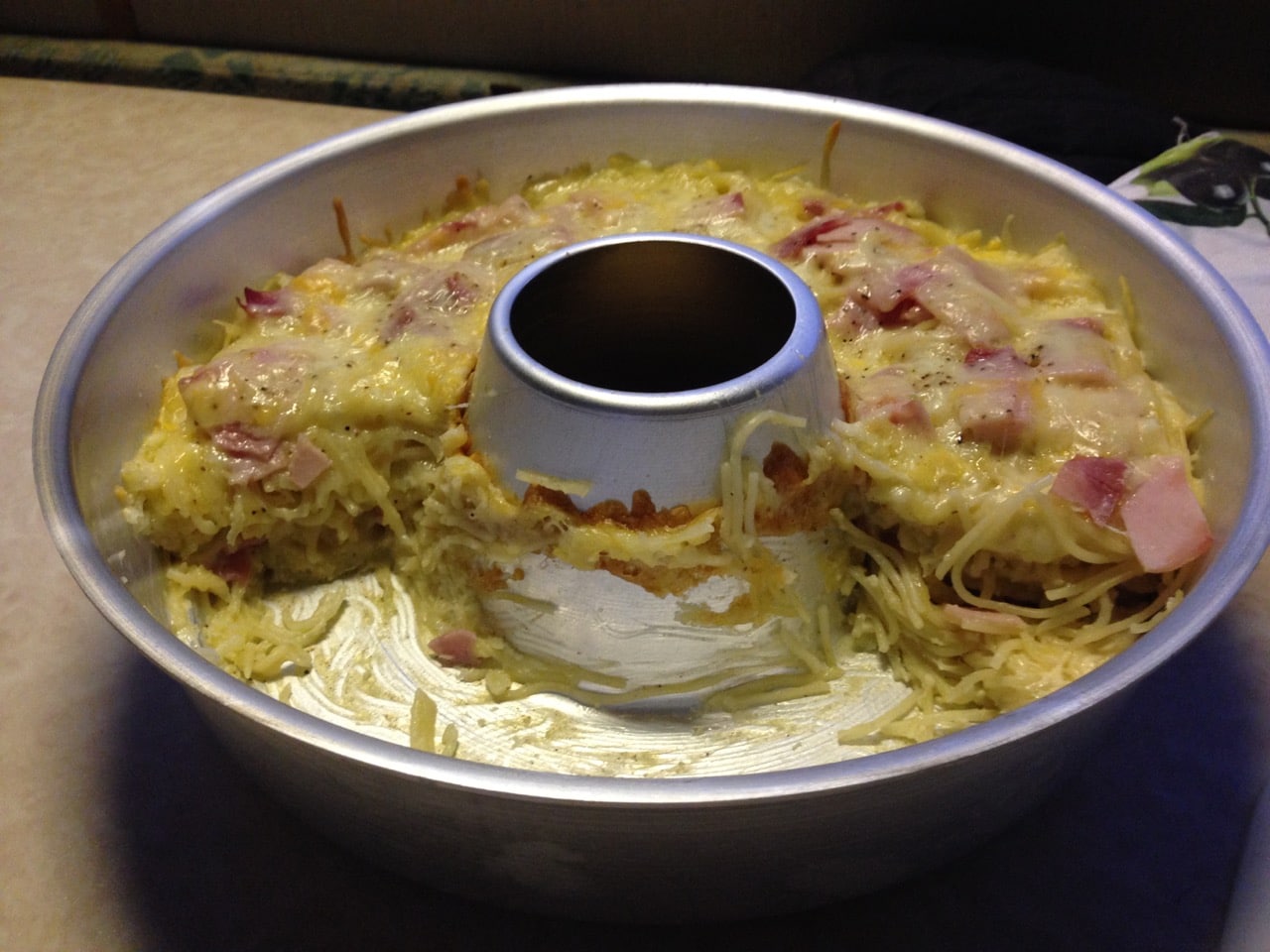 Ein wie eine Kuchenform aussehender Omnia-Camping-Backofen halb gefüllt mit einem Auflauf aus Spaghetti, Schinken und Käse