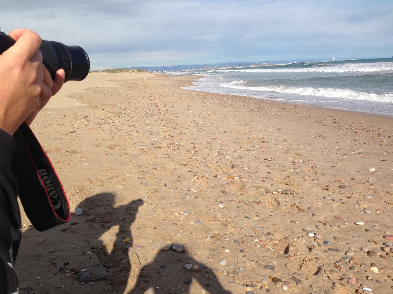 Links im Bild ein Fotoapparat und die Schatten von zwei fotografierenden Menschen, recht Strand und Meer