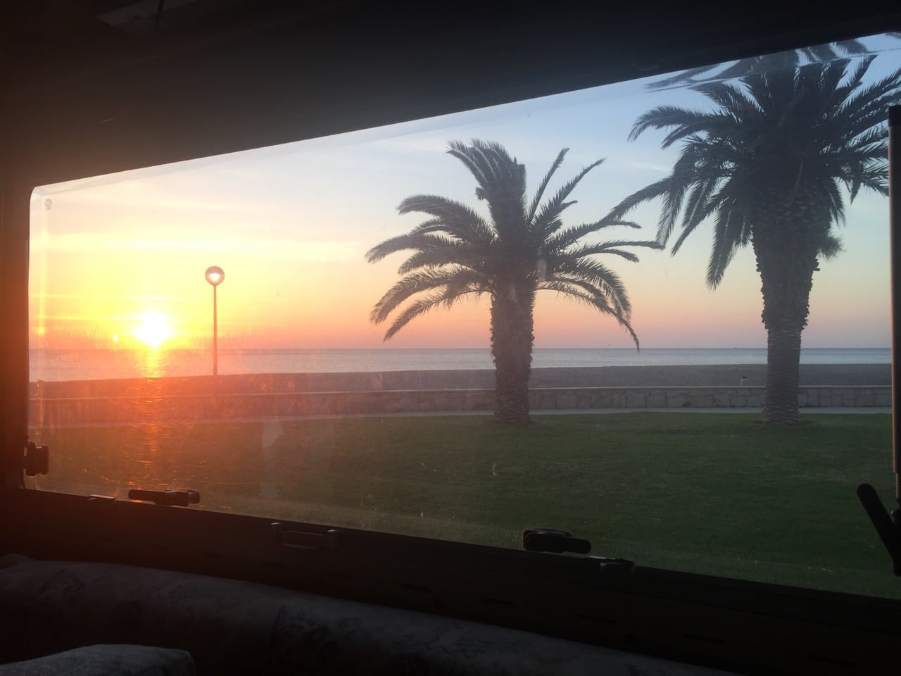 Blick aus einem Wohnmobilfenster auf das Meer, Palmen und die aufgehende Sonne