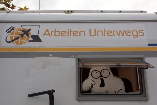 Ein Wohnmobil von hinten, aus dem Fenster schaut ein Kissen in Katzenform und über dem Fenster ein Aufkleber mit "Arbeiten unterwegs"