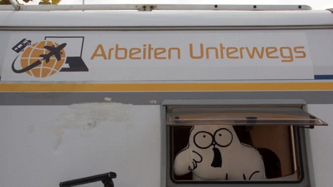 Ein Wohnmobil von hinten, aus dem Fenster schaut ein Kissen in Katzenform und über dem Fenster ein Aufkleber mit "Arbeiten unterwegs"