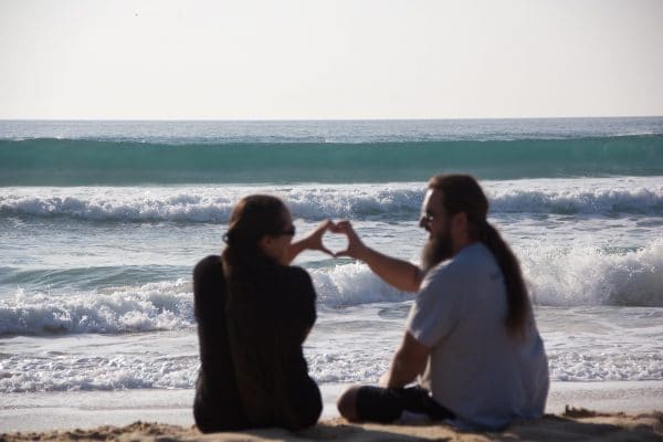 Ein Paar sitzt am Strand, formt mit den Händen ein Herz, im Hintergrund das Meer