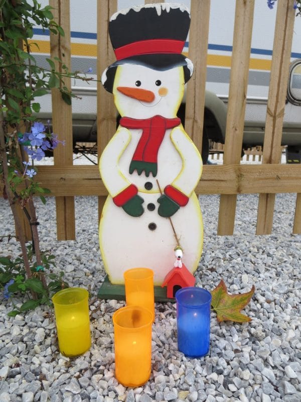 Ein Schneemann aus Holz vor einem Holzzaun auf hellem Kies, davor vier Windlichter