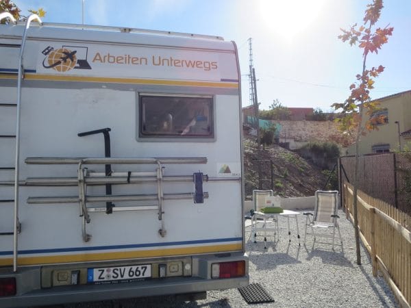 Ein Wohnmobil von hinten daneben Stühle und ein Tisch und Sonne von vorne