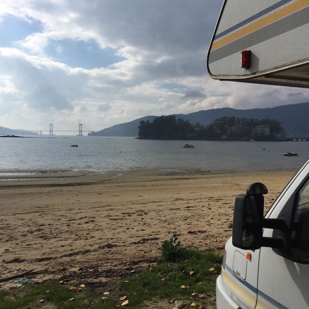 Das Fahrerhaus von einem Wohnmobil, davor ein Strand, Wasser und im Hintergrund eine Brücke